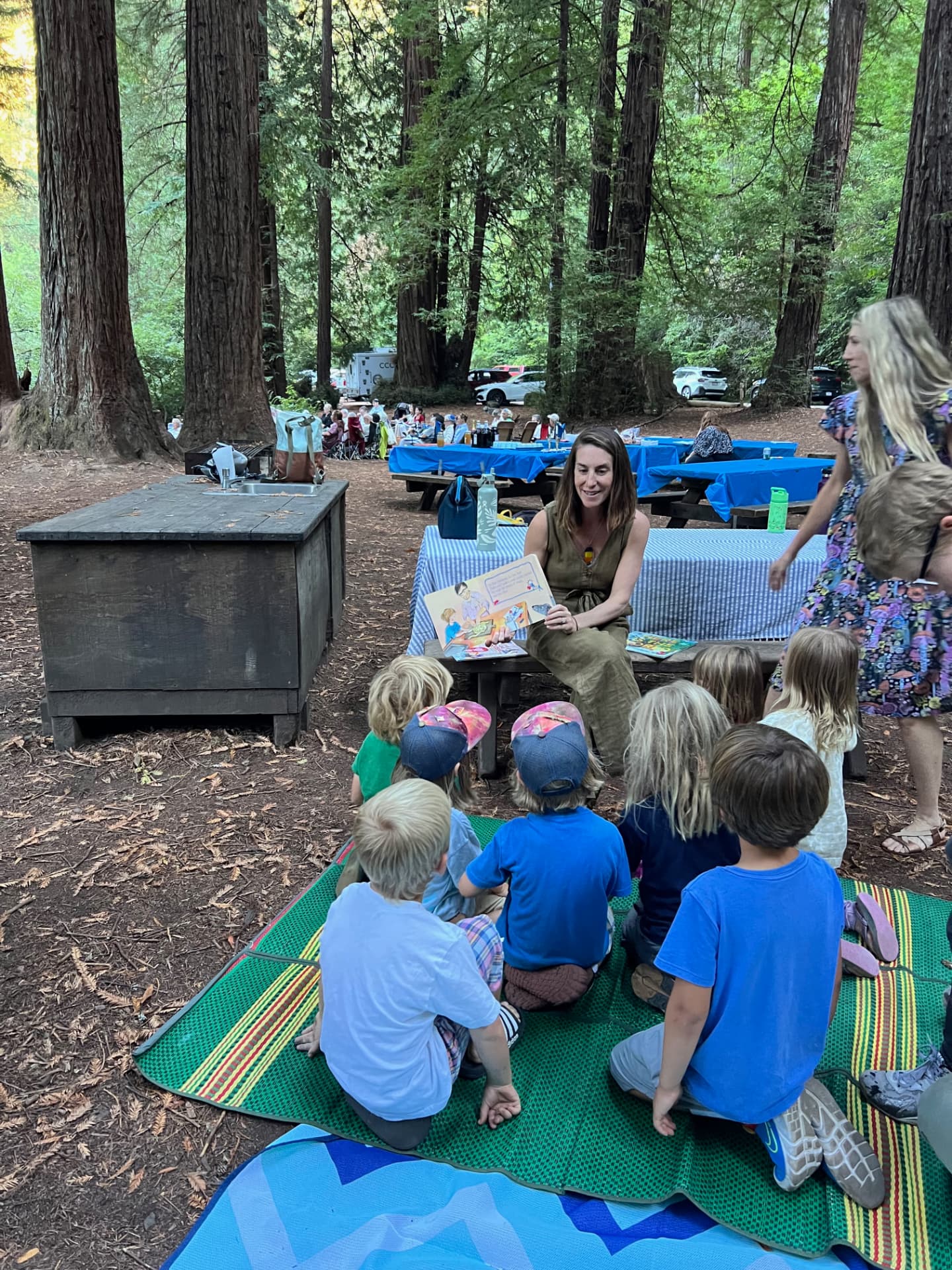 A teacher reads to a circle of children outdoors among redwood trees